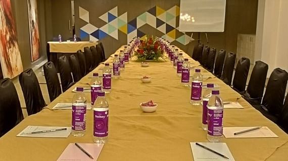 A long conference table set up for a meeting. There are water bottles, notepads, and pens arranged neatly at each seat. A whiteboard stands in the background, and the walls are decorated with a geometric pattern.